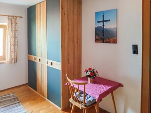 A room with a wooden floor, a wooden chair with a cushion, a table with a pink cloth, a cross painting, and a window with curtains.