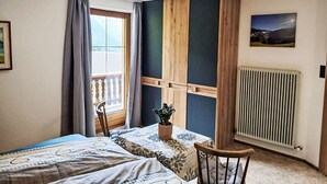 Un dormitorio con una cama, una silla, una planta en maceta, una ventana con cortinas, un radiador y un cuadro colgado en la pared.