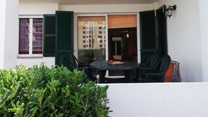 Un balcón con una mesa y sillas, una ventana con persianas verdes, y una vista de un edificio al fondo.