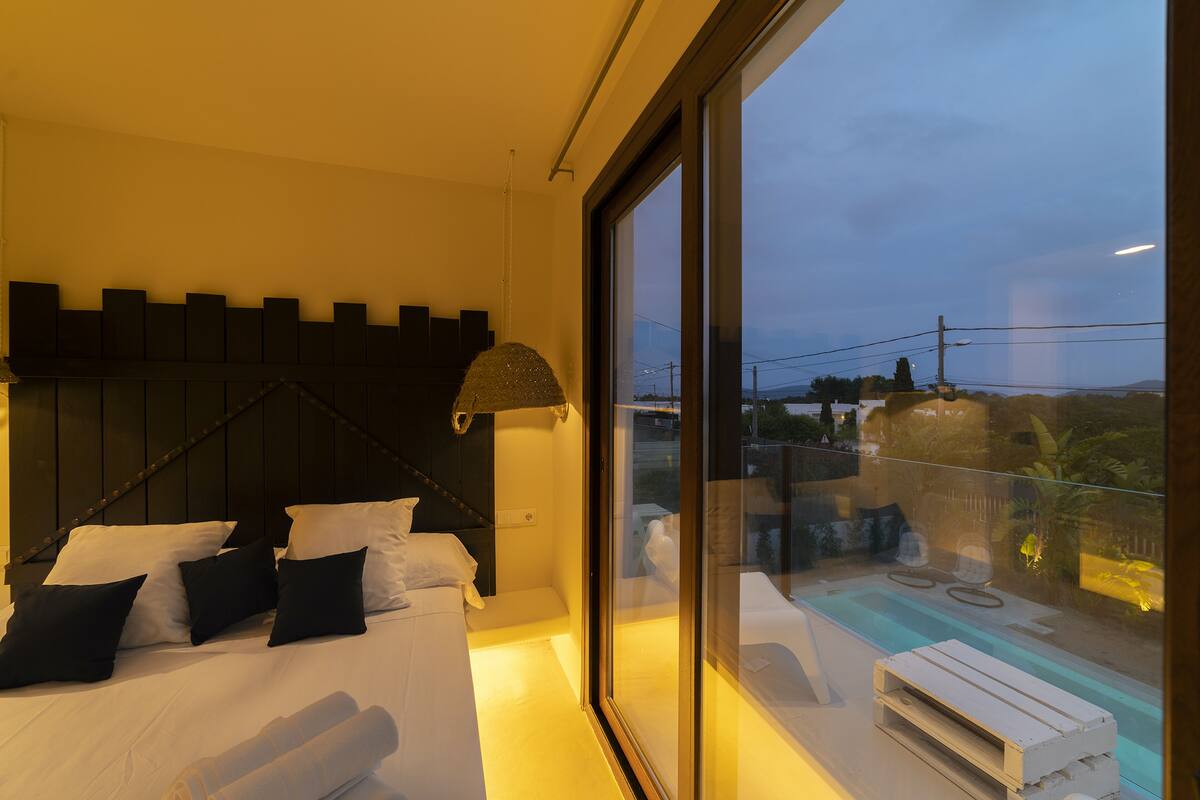 Un dormitorio con cama, almohadas, cabecera, ventana y vistas al exterior.