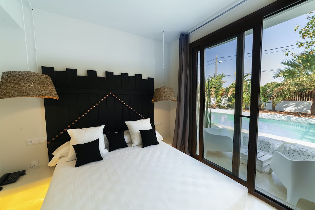 Habitación de hotel con una cama, cabecera negra, dos lámparas y vista a una piscina a través de ventanas amplias.