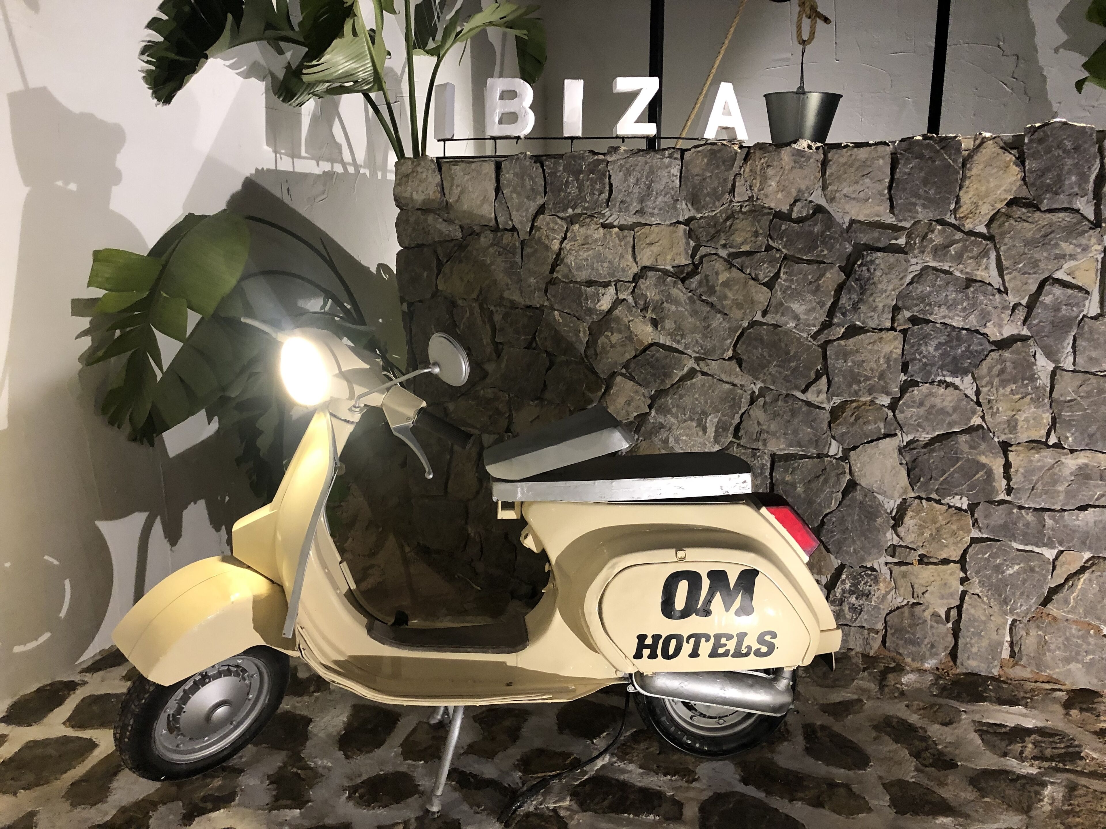 A vintage yellow scooter parked in front of a stone wall with the sign "OM HOTELS" and a potted plant to the left.