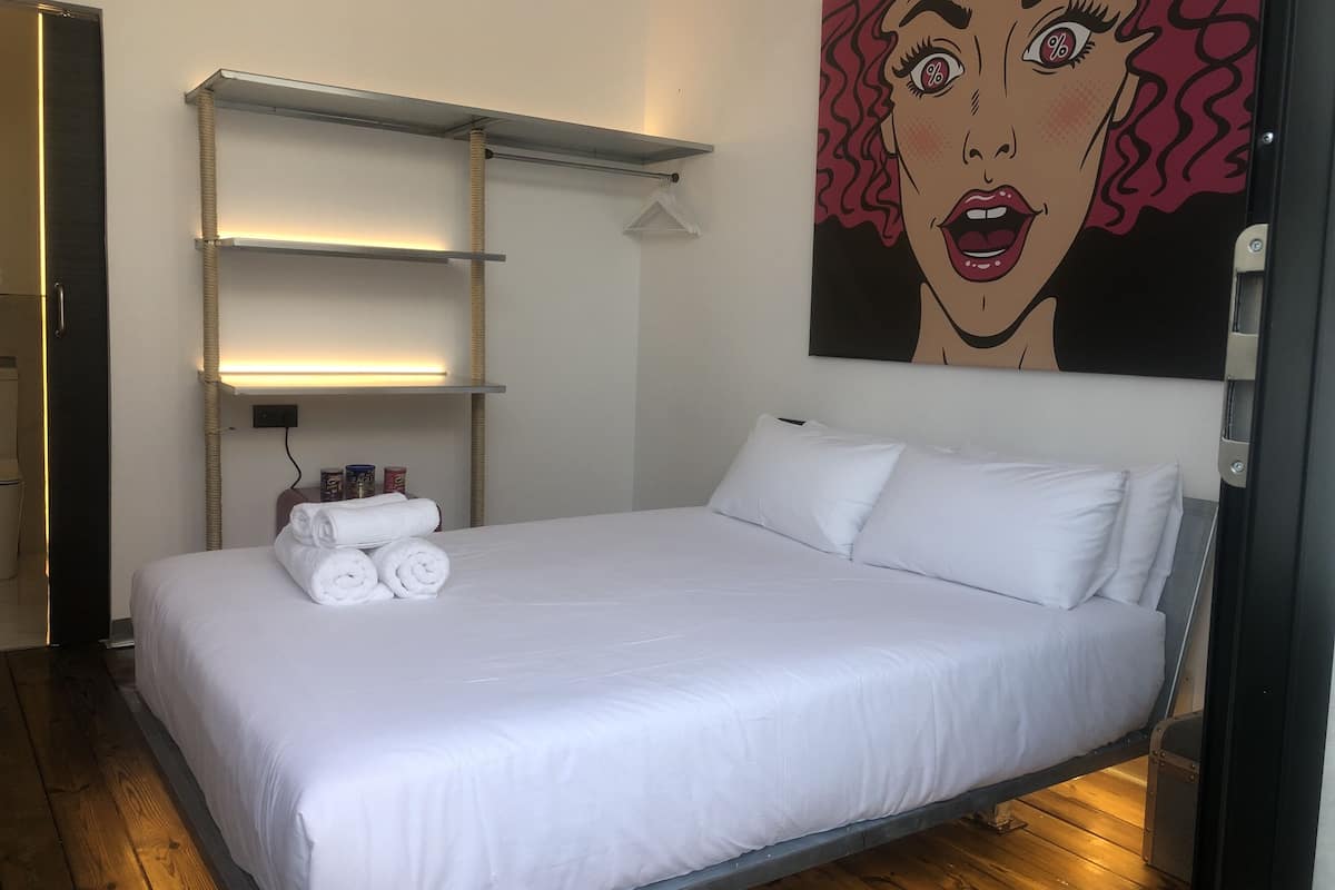 Una cama bien hecha con ropa blanca y almohadas, un estante con una lámpara y un cuadro de gran formato de arte pop en la pared.