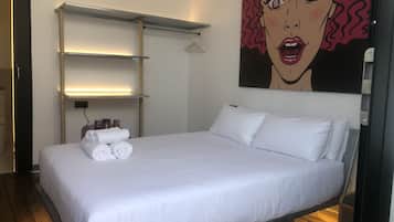 Una cama bien hecha con ropa blanca y almohadas, un estante con una lámpara y un cuadro de gran formato de arte pop en la pared.