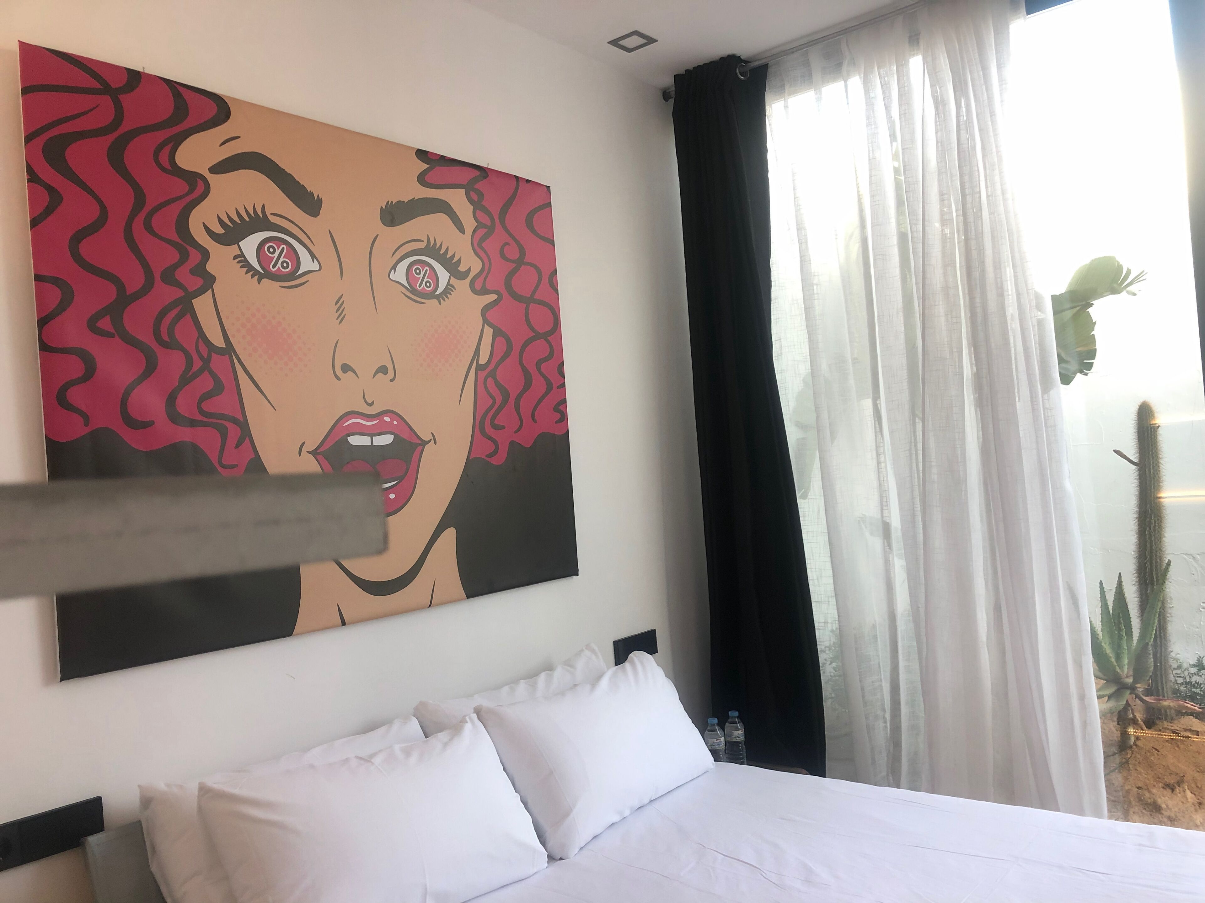 Quarto de hotel com cama, almofadas brancas, uma pintura grande na parede e uma janela com cortinas finas.