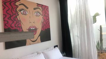 Quarto de hotel com cama, almofadas brancas, uma pintura grande na parede e uma janela com cortinas finas.