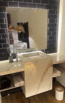 Un baño con lavamanos de mármol, un espejo y pared revestida de baldosas negras.