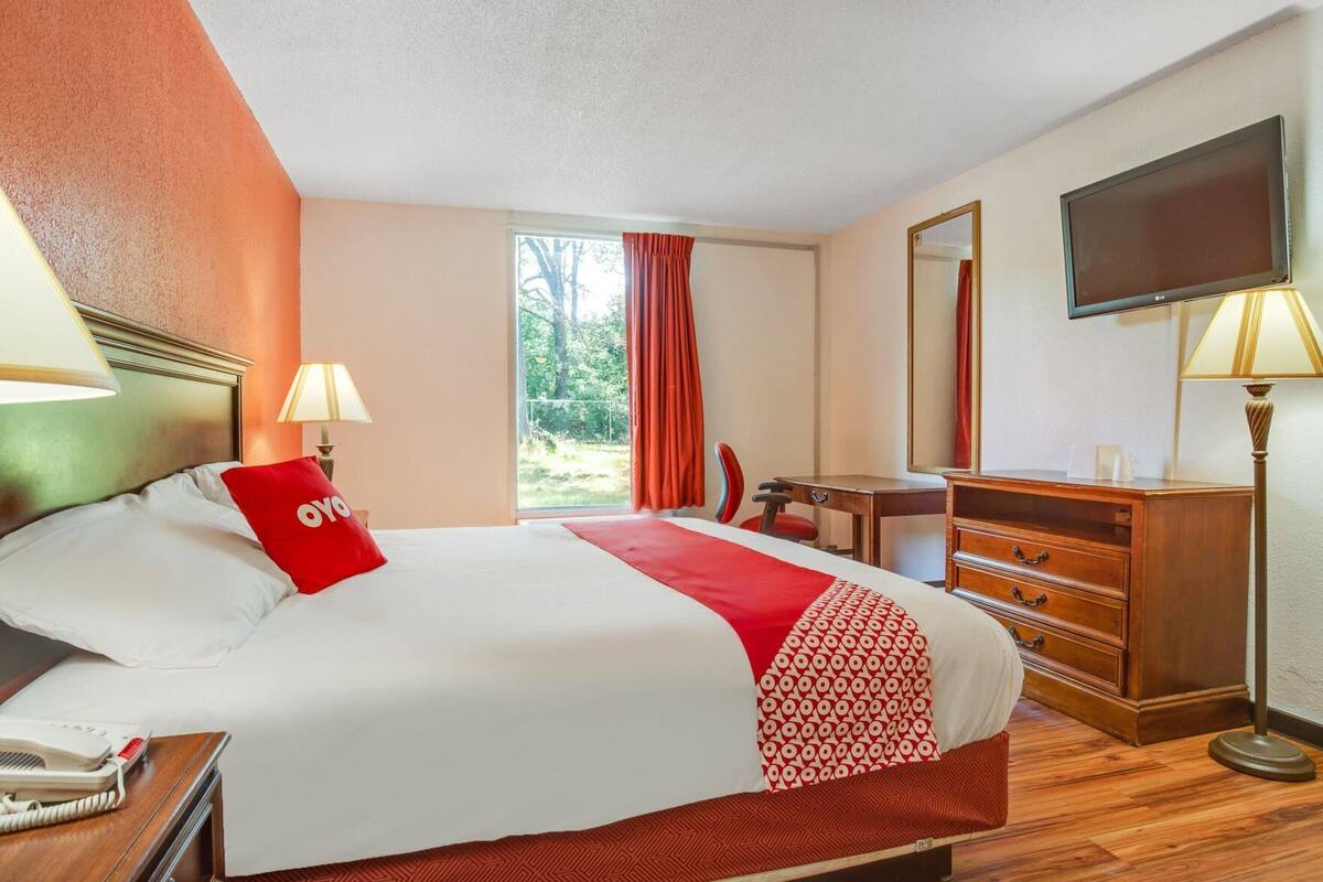 A hotel room with a bed, bedside table, desk, dresser, mirror, TV, and a window with curtains.