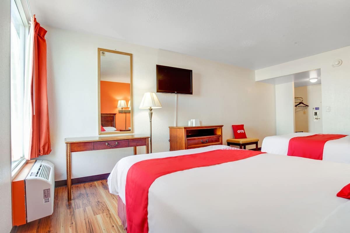 A hotel room with two beds, a desk, a mirror, a TV, and a heater.