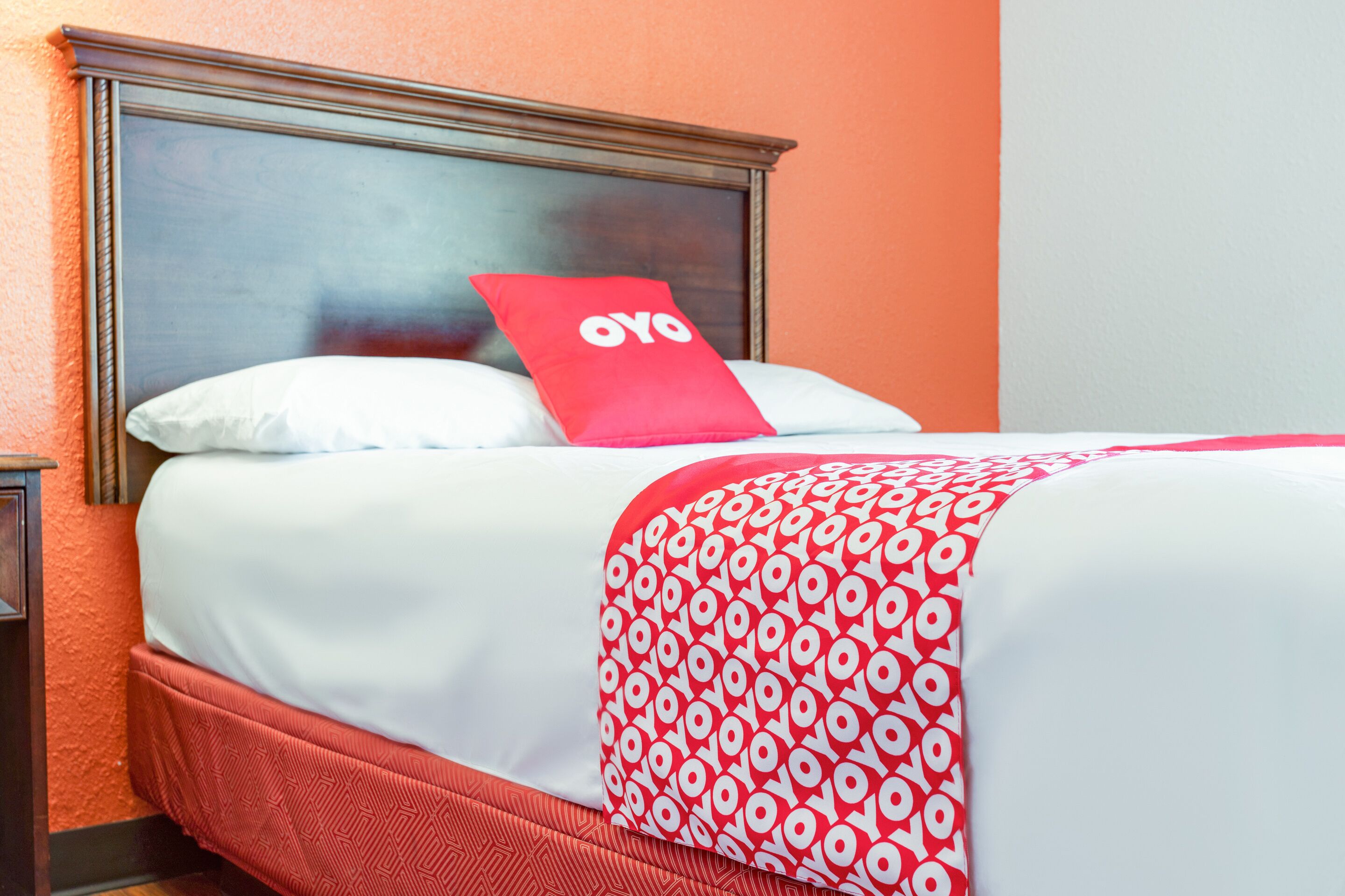 Habitación de hotel con una cama, una colcha de diseño rojo y blanco, un almohadón con el logotipo 'oyo' y un cabecero de madera.