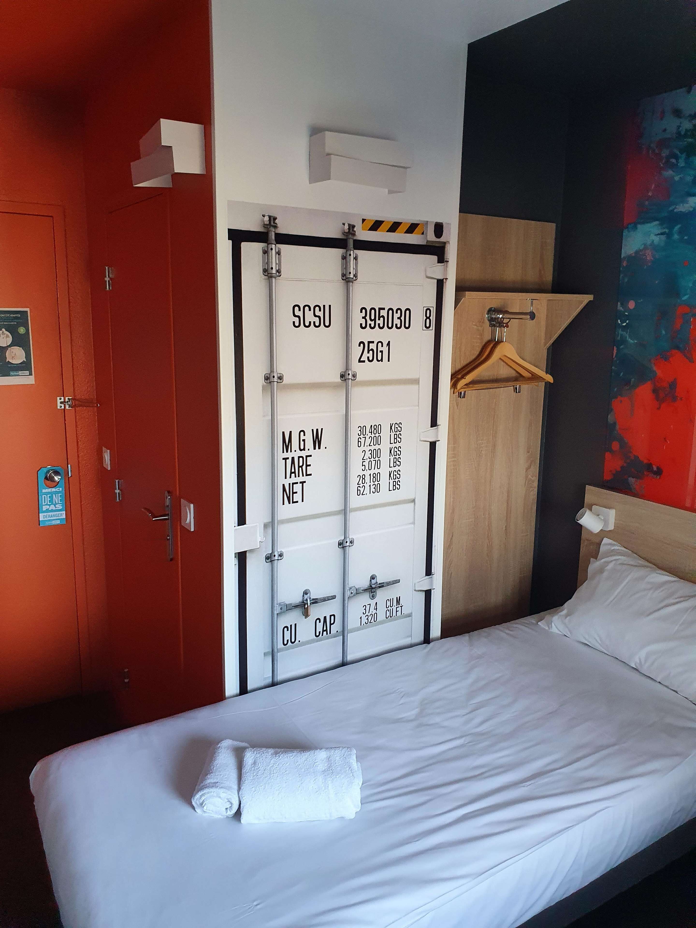 Une chambre d’hôtel avec un lit, une porte rouge, un mur blanc orné d’un tableau d’affichage et une peinture abstraite aux couleurs vives.