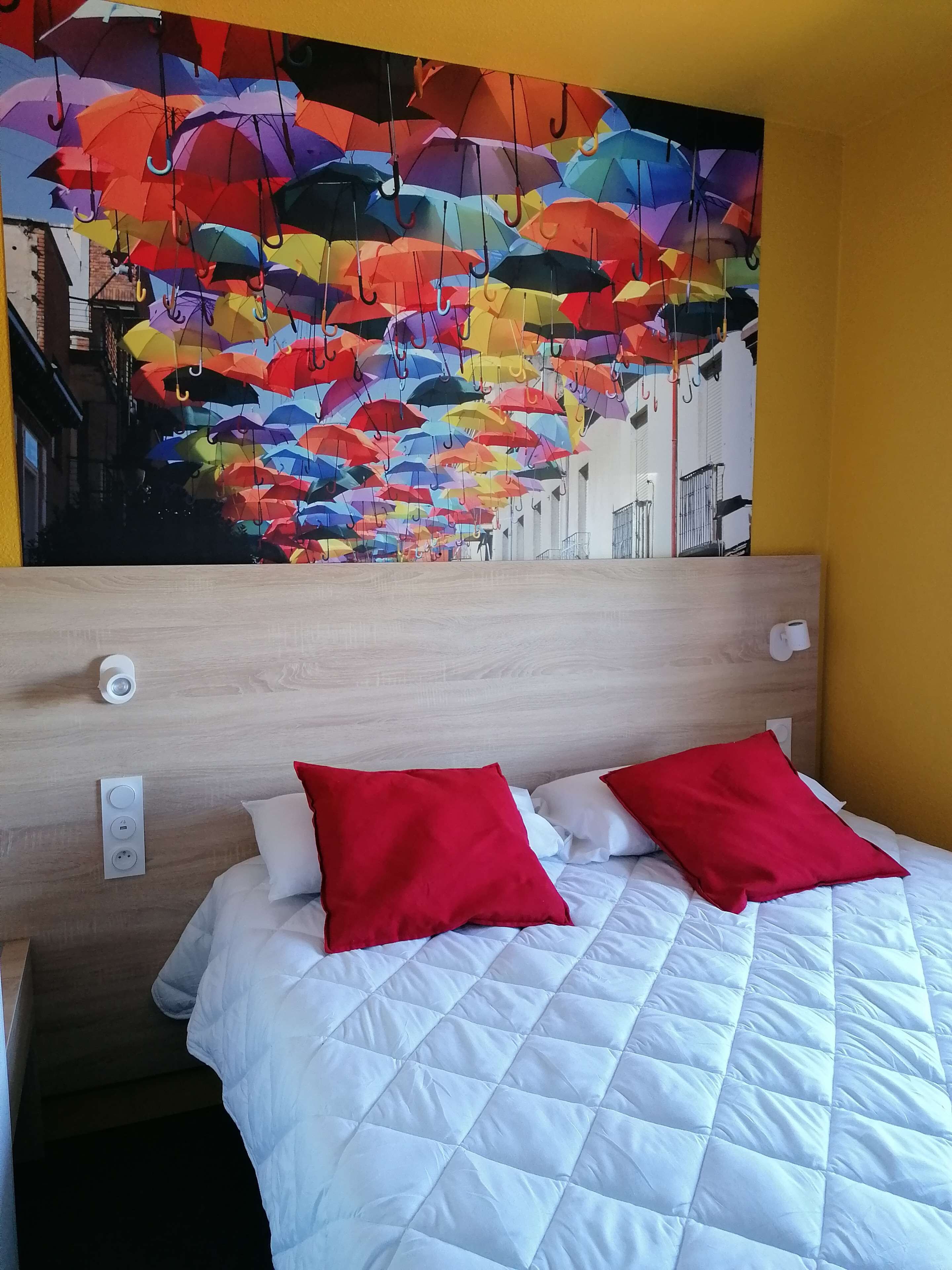 Une chambre d’hôtel avec un lit, deux oreillers rouges, une couette blanche matelassée, une tête de lit en bois et une fresque murale représentant des parapluies colorés.