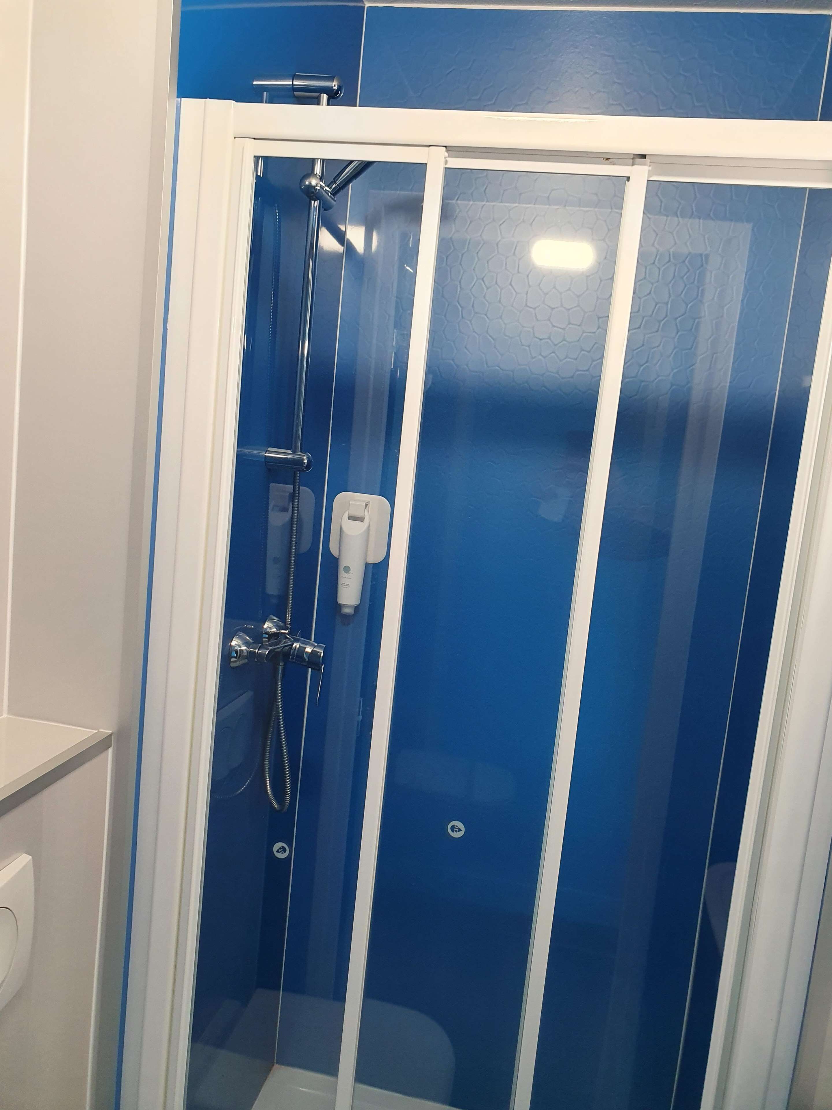 Une cabine de douche fermée par des parois en verre, dotée d’une porte blanche, d’un pommeau de douche à main et d’un distributeur de savon à main.