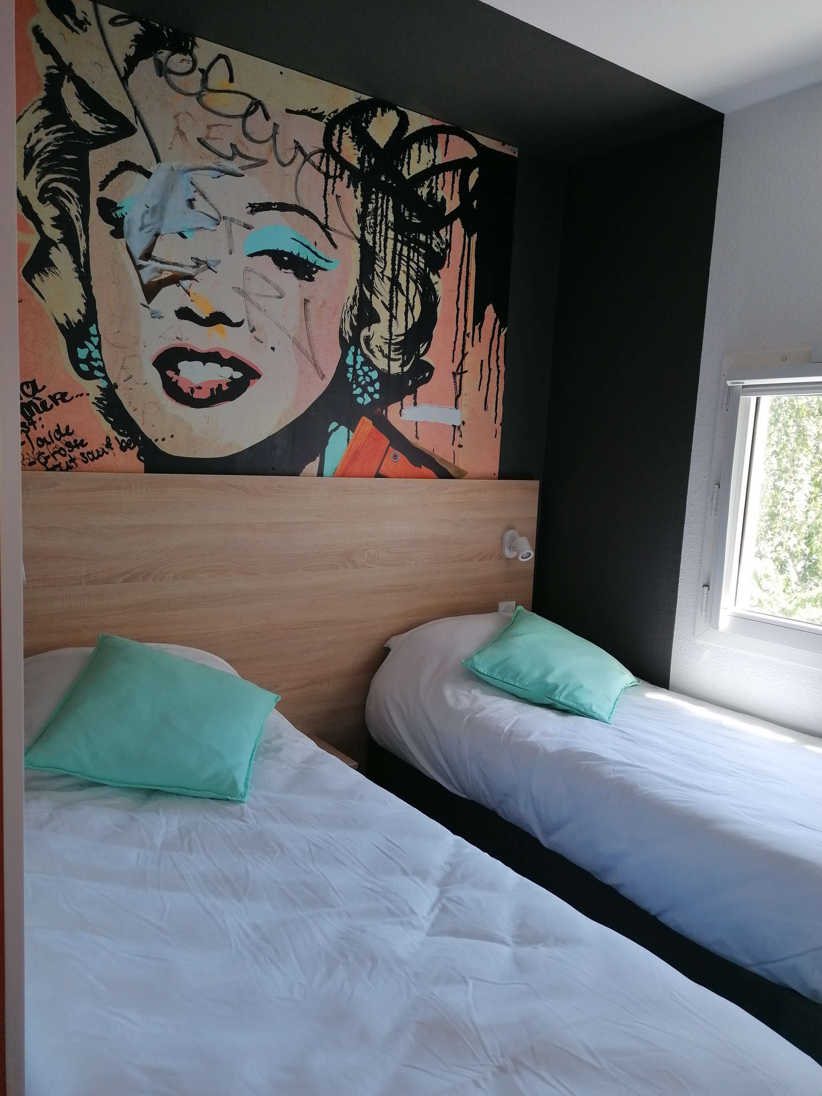 Une chambre d’hôtel avec deux lits, chacun agrémenté de draps blancs et d’oreillers turquoise. Une grande fresque colorée orne le mur au-dessus des lits.