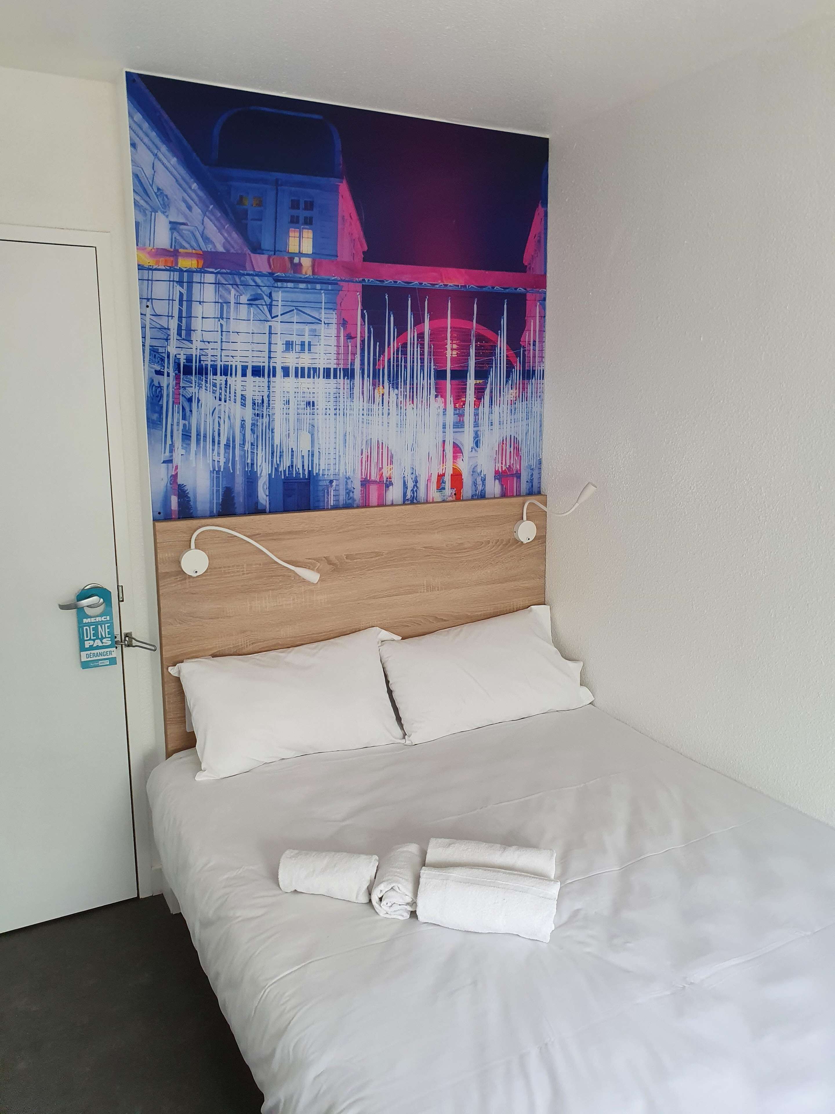 Une chambre d’hôtel avec un lit, du linge de lit blanc, une tête de lit en bois et un grand tableau abstrait accroché au mur.