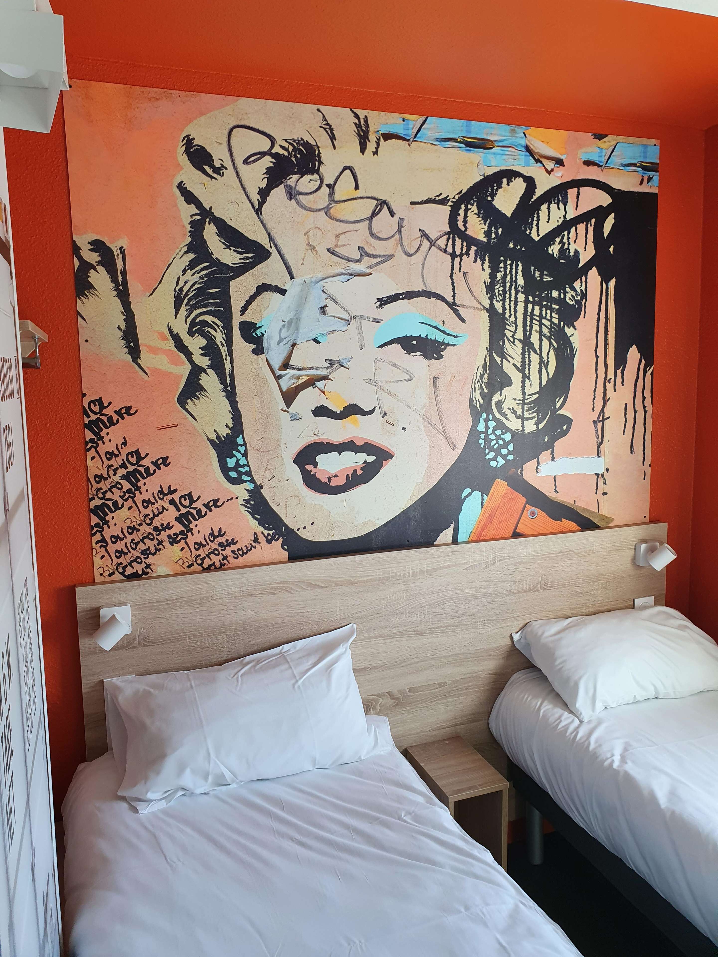 Une chambre d’hôtel avec une grande fresque murale, deux lits avec du linge de lit blanc et une tête de lit en bois.