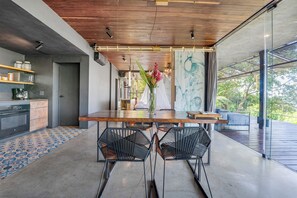 A modern kitchen with a wooden ceiling, a dining table with chairs, and a view of greenery outside. - Atelier NEW design creation at Art Villas (Uvita)