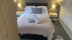 A neatly made bed with white linens and a folded towel, two bedside lamps, and a small nightstand with a phone. - Heatherland Cottage (Skeabost)