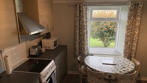 A compact kitchen with a stove, microwave, and toaster. There's a dining table with chairs and a window with curtains. - Heatherland Cottage (Skeabost)