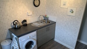 A compact kitchen with a washing machine, a kettle, and a sink. - Heatherland Cottage (Skeabost)