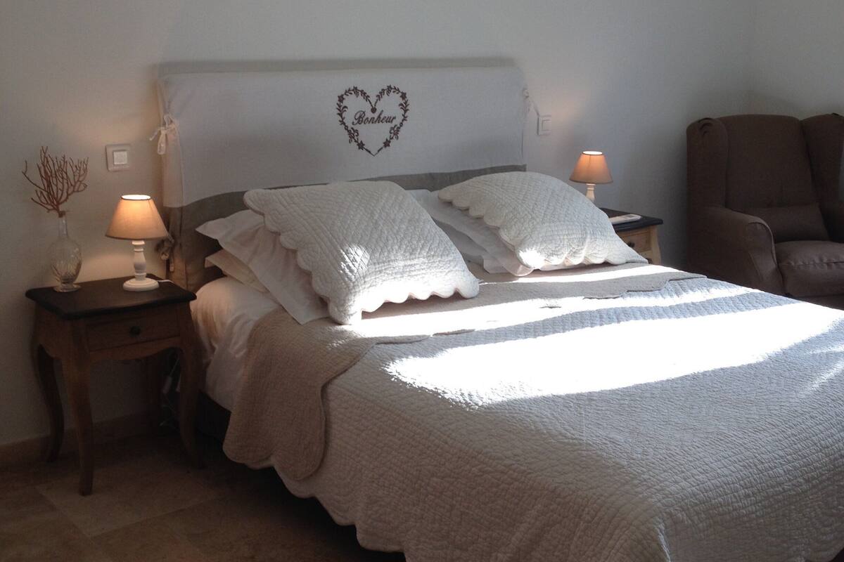 Un lit bien fait, agrémenté d’une décoration en forme de cœur sur la tête de lit, deux lampes de chevet et une chaise placée dans un coin.