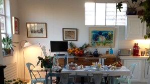 Un coin repas avec un sol carrelé, une table dressée pour quatre personnes et des chaises. On y trouve des plantes en pot, un tableau accroché au mur et un téléviseur posé sur un meuble.