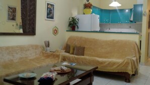 A living room with a sofa, a coffee table, and a kitchen area with cabinets and a refrigerator. - Palinas comfy home!! (Kalamata)