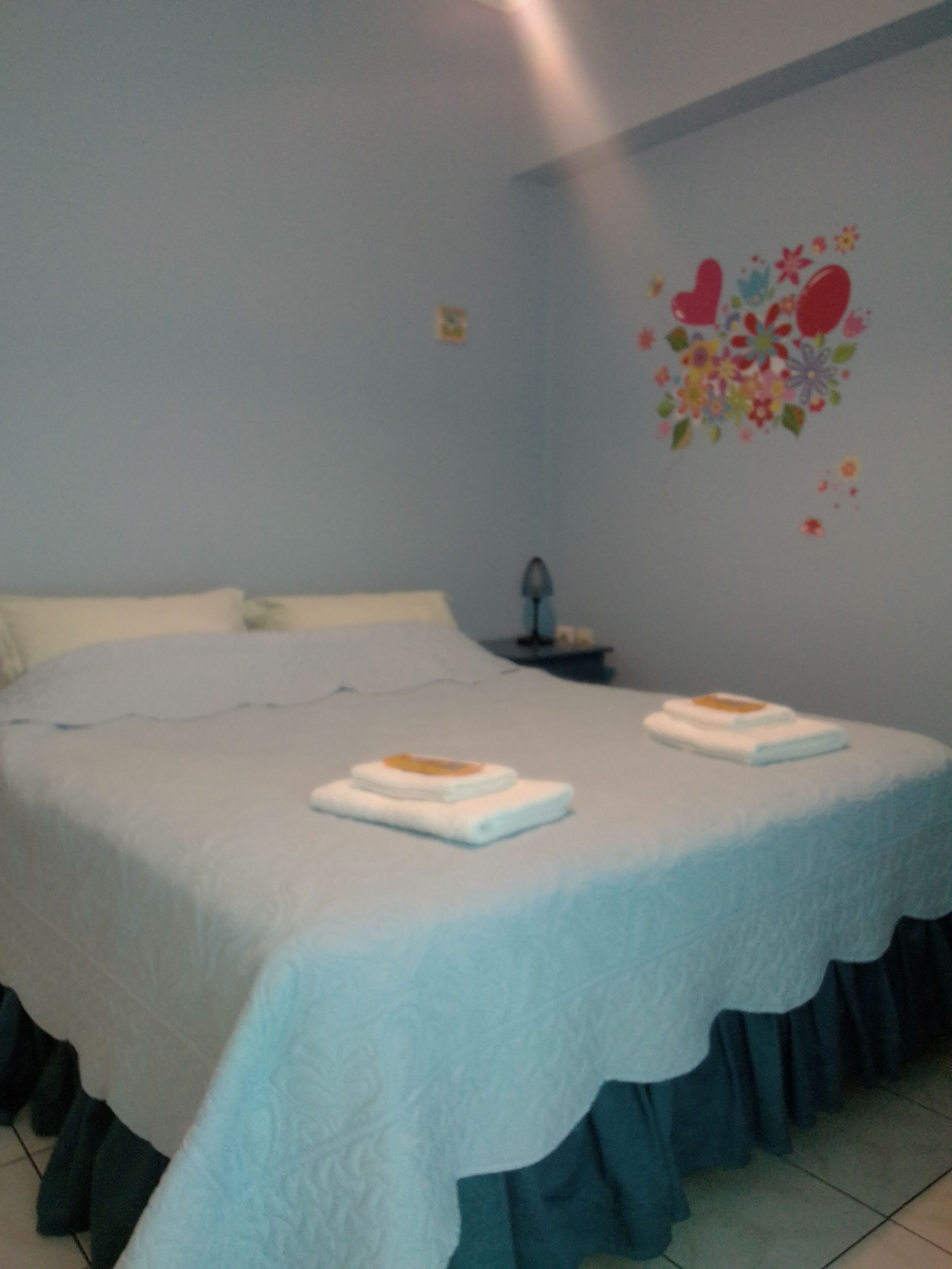A double bed with a blue bedspread and a ruffled skirt, two white towels, and a nightstand with a lamp.