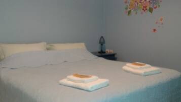 A double bed with a blue bedspread and a ruffled skirt, two white towels, and a nightstand with a lamp.