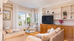 A living room with beige sofas, a coffee table, a TV on a wooden cabinet, and large windows with curtains.