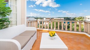 A balcony with a white sofa and a small table with flowers, overlooking a cityscape with buildings and mountains.