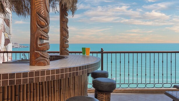 Une terrasse circulaire en bois offrant une vue sur la mer, dotée d’une rambarde en métal et d’un mur en pierre.