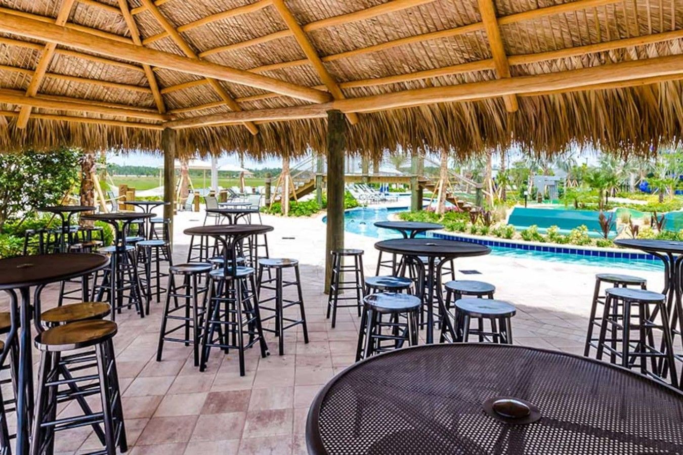 An outdoor patio area with tables and stools, a pool, and a view of a tropical landscape.