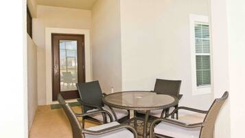 A small balcony with a glass table and wicker chairs, a door, and a window with blinds.