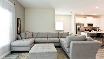 A modern living room with a sectional sofa, ottoman, and a kitchen in the background.