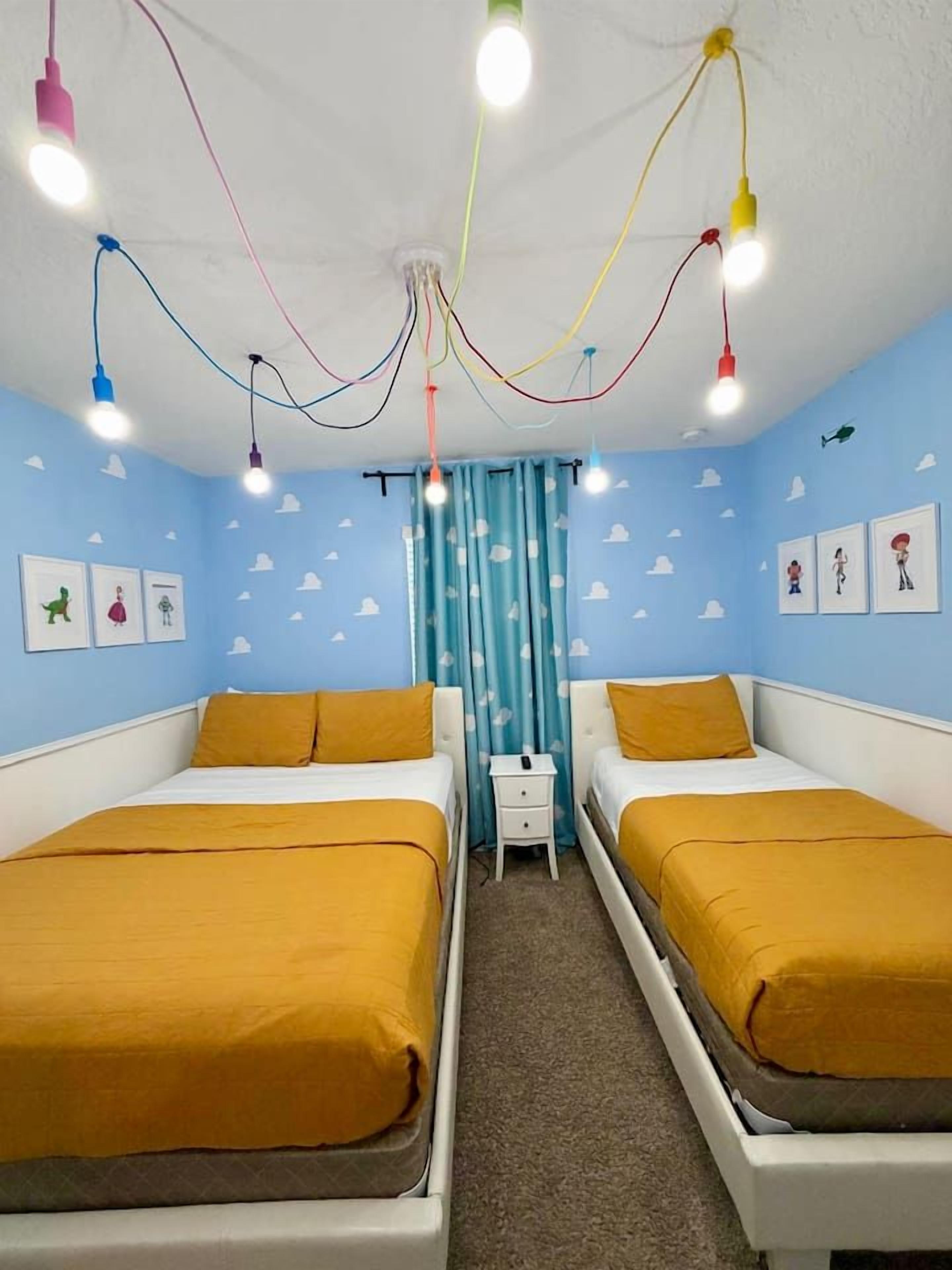 Una habitación con dos camas, una mesita de noche e iluminación colgante de colores en el techo.