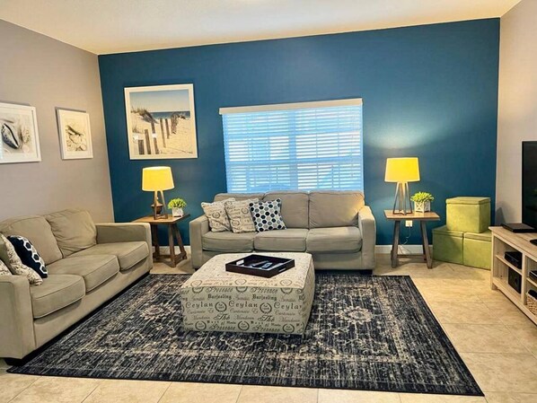 A living room with a sofa, two side tables with lamps, a coffee table with a tray, and a large window with blinds.