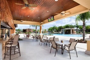 A covered outdoor dining area with tables and chairs, a pool, and a waterfall feature. - Resort Home Sleeps 12 w/private Pool! (Kissimmee)