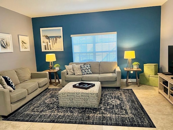 A living room with a beige sofa, a matching ottoman, a blue accent wall, and a large window with blinds.