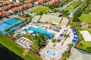 An aerial view of a resort with a large swimming pool, multiple lounge areas, and a well-maintained lawn.