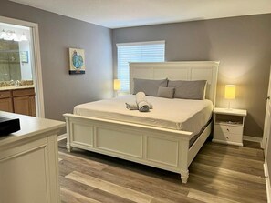 A bedroom with a large bed, bedside tables, a nightstand, a dresser, a window with blinds, and a painting on the wall.
