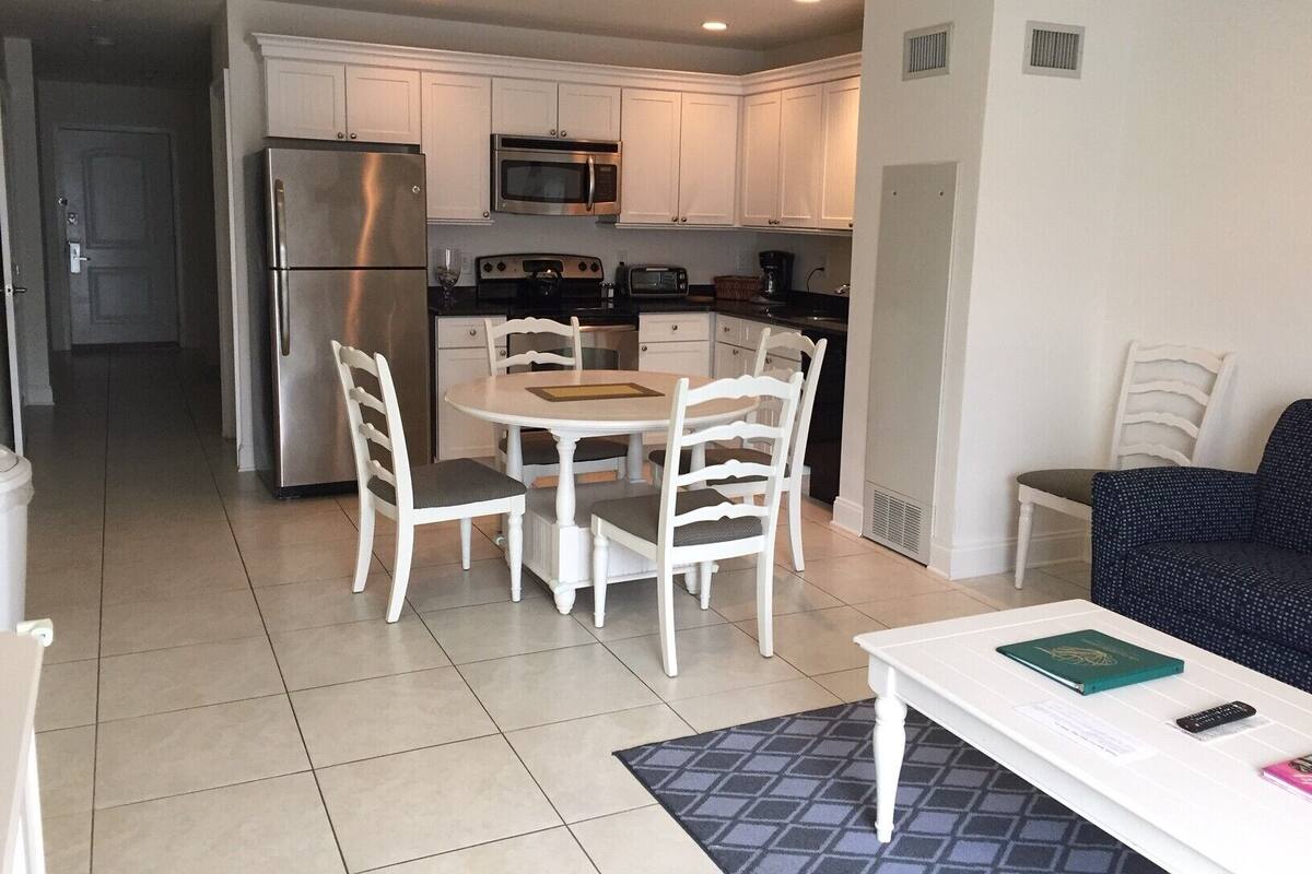 A compact kitchen with white cabinetry, a stainless steel refrigerator, a microwave, and a stove. There is a dining table with four chairs, and a small white table with a book and remote control.