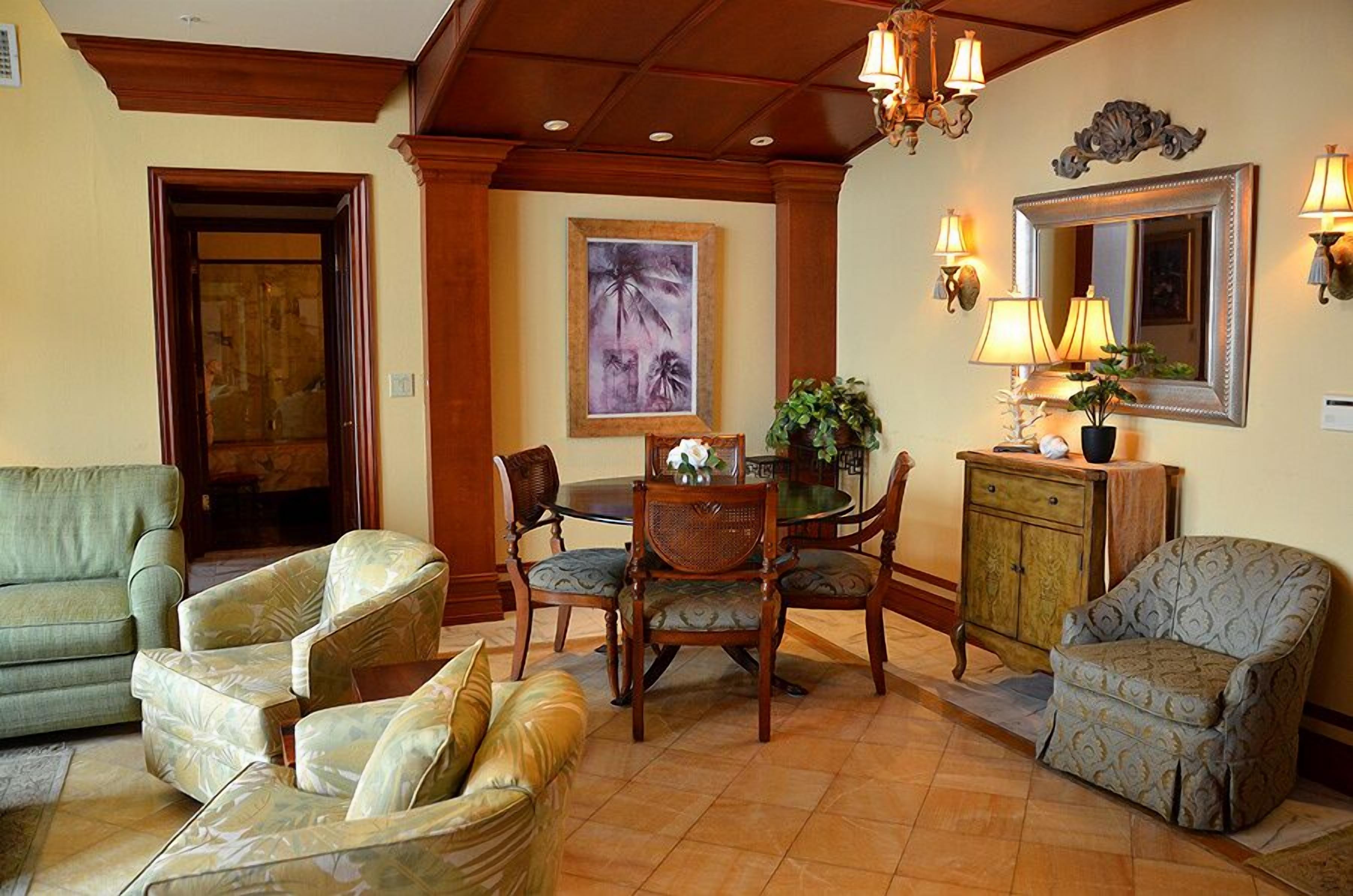 A living room with a green sofa, patterned armchairs, a dining table with chairs, a cabinet, and a large mirror with wall sconces.
