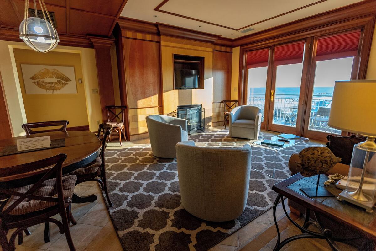 A hotel room with a dining area, two armchairs, a sofa, a TV, and a view of the ocean.
