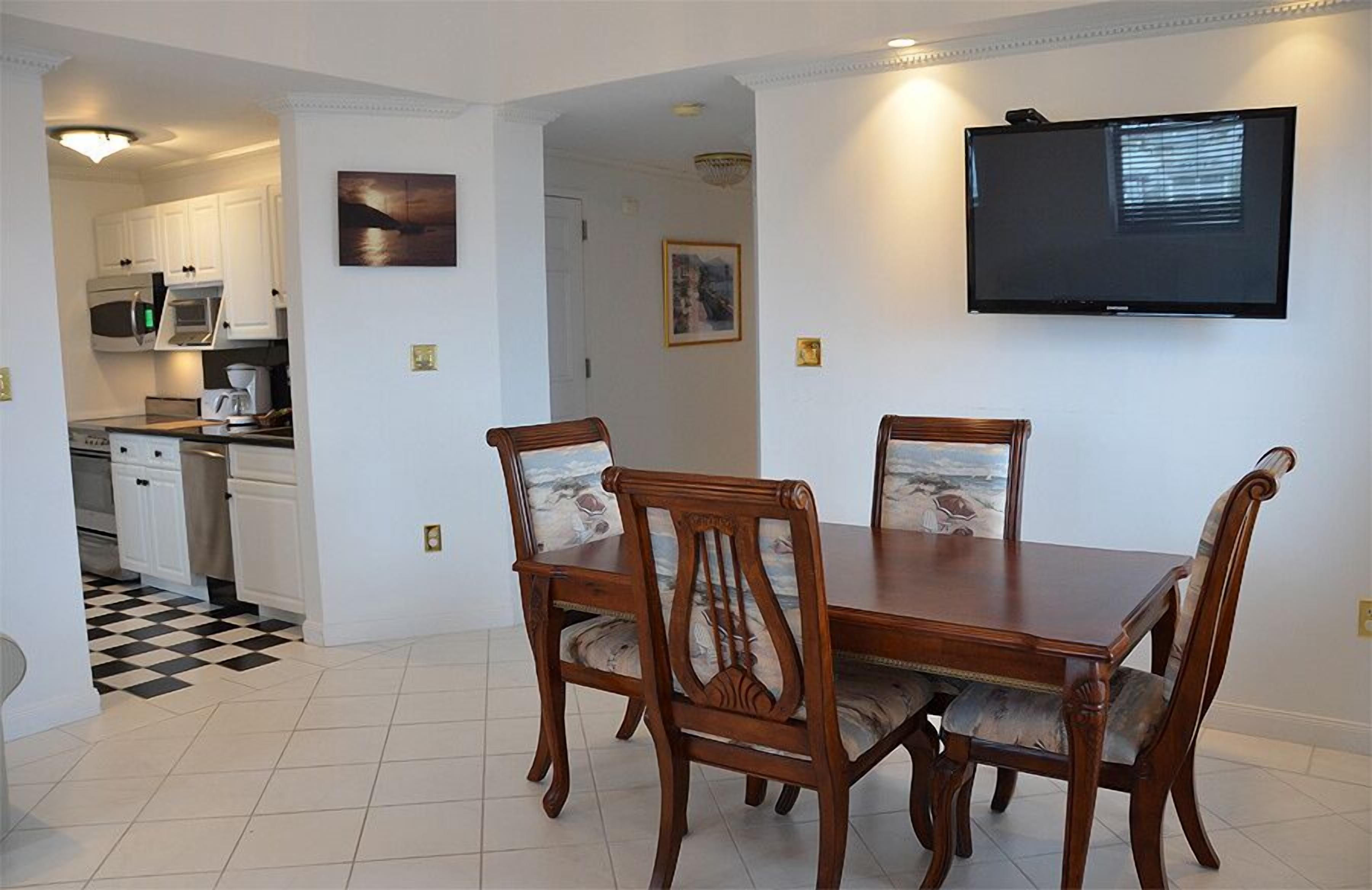 Un comedor con mesa y sillas de madera, una cocina al fondo y un televisor montado en la pared.
