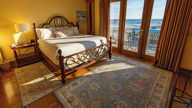 Ein Schlafzimmer mit einem großen Bett, einem Nachttisch, einer Lampe, Blick auf den Strand und einem Balkon mit Sitzgelegenheiten.