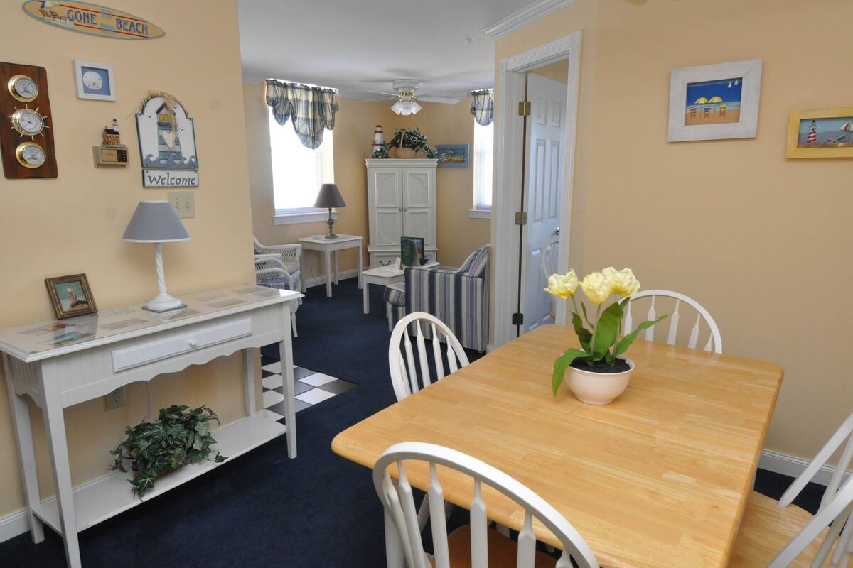 A dining area with a wooden table, white chairs, a potted plant, and a wall clock.