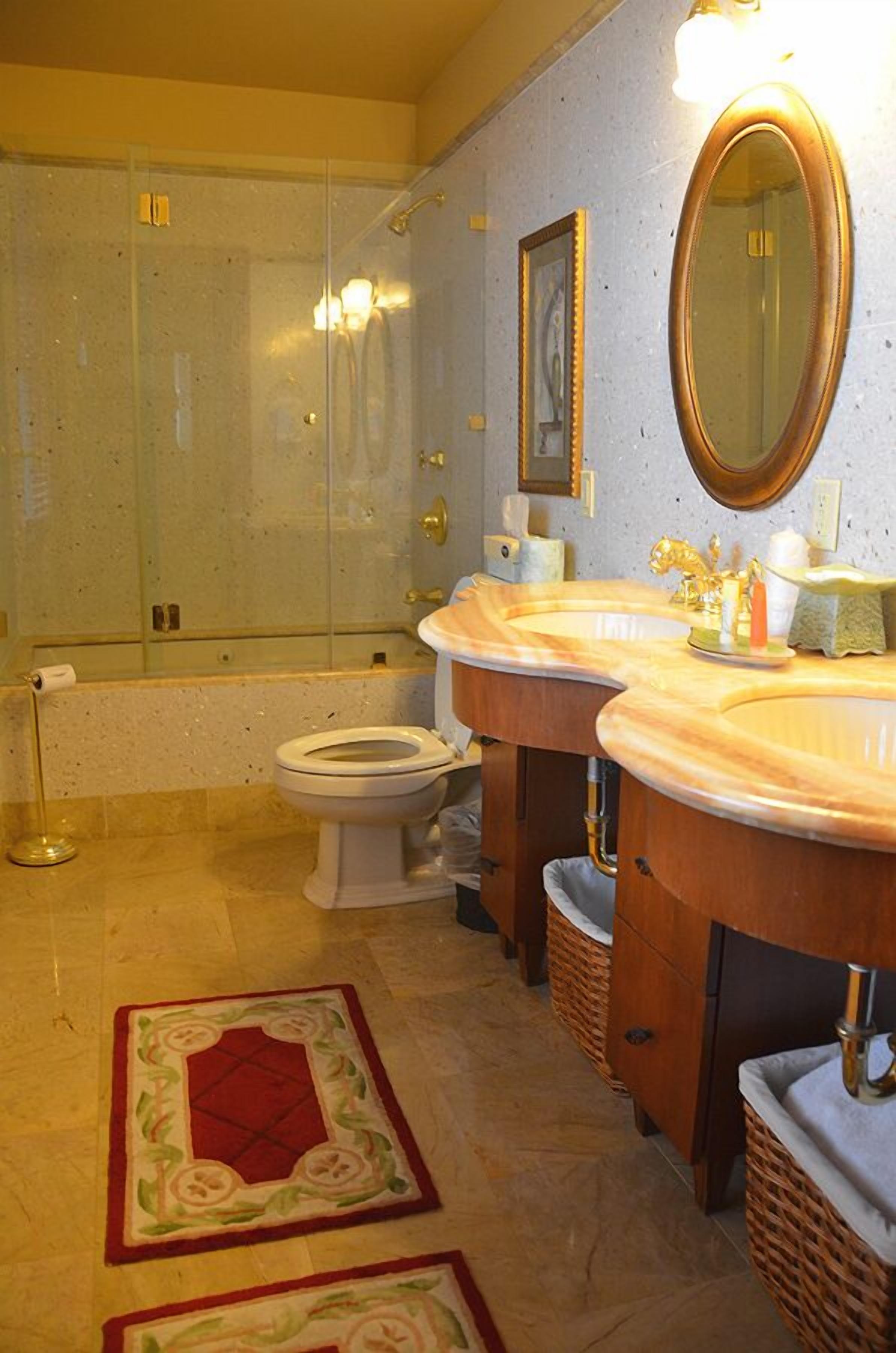 A bathroom with a glass-enclosed shower, two sinks with gold fixtures, a large mirror, and a red decorative rug.
