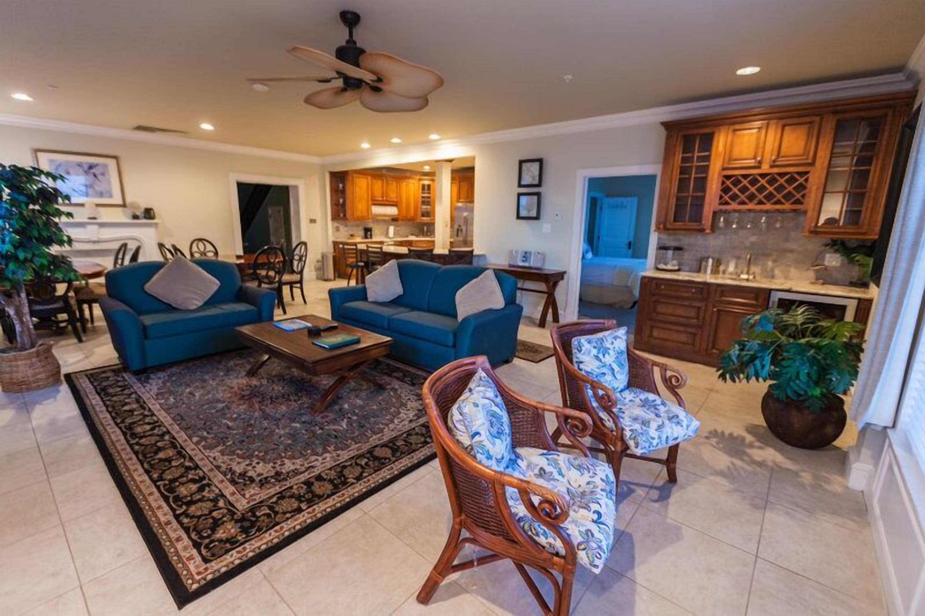 A spacious living room with a blue sofa set, a coffee table, and wicker chairs. A kitchen with wooden cabinets is visible in the background.