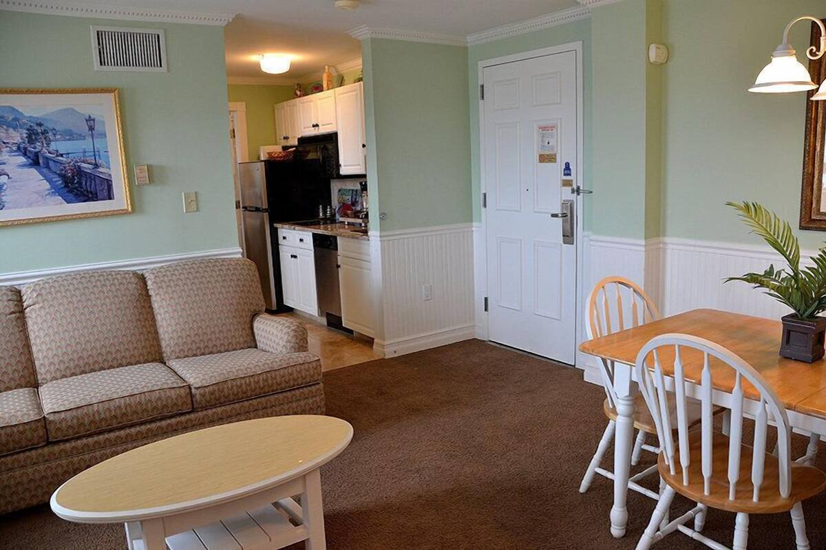 A living room with a sofa, a dining table with chairs, a kitchen in the background, and a door.